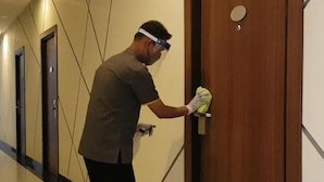 A person cleaning a wooden door in a hallway with a patterned wall and a door with a round handle. - Kawana Hotel (Padang)