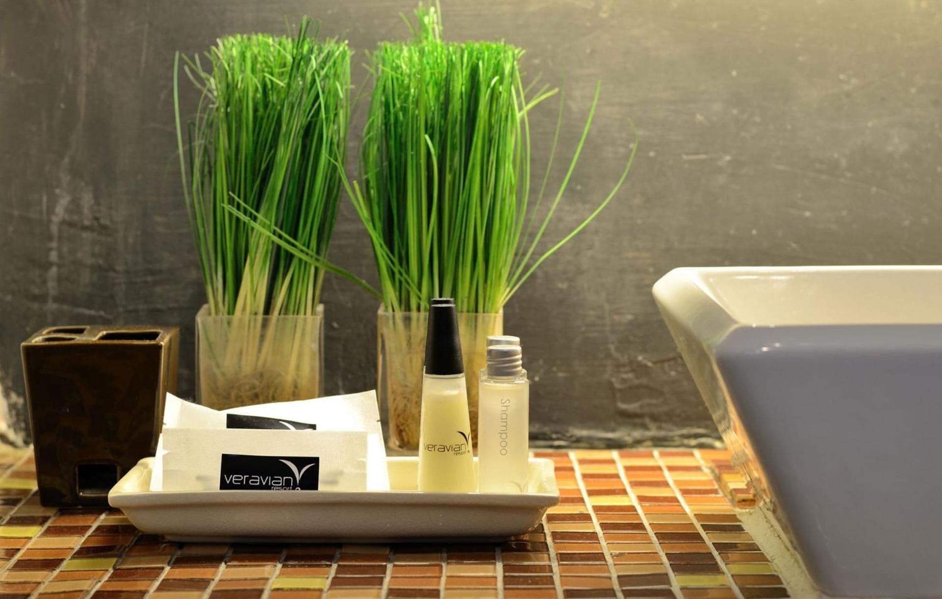 A bathroom with a tiled floor, a white sink, and a bathtub. There are two glass containers with green grass and two bottles of shampoo.