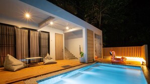 Un espace piscine extérieur moderne, doté d’une terrasse en bois, d’un coin salon avec des coussins et d’un flotteur décoratif en forme de flamant rose.