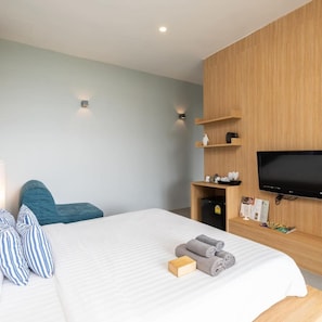 Moderni hotellihuone, jossa on sänky, televisio, puinen hylly, jossa on hygieniatarvikkeita, ja pieni jääkaappi.