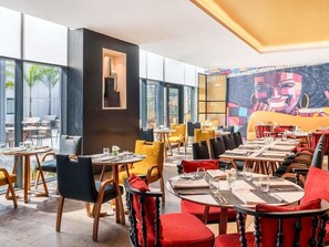 En modern restaurang med runda bord och stolar, stora fönster och en färgstark väggmålning.