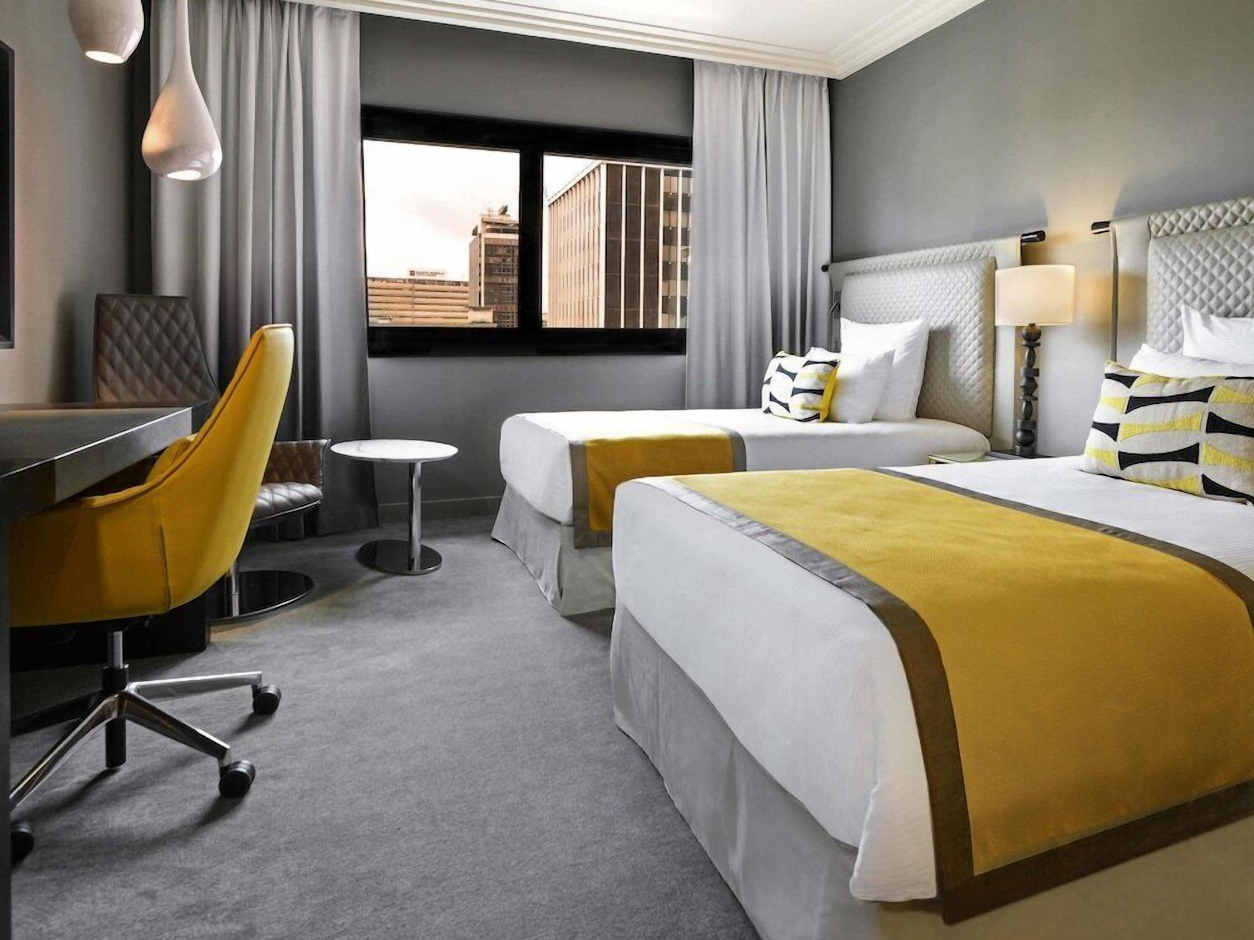 A hotel room with two beds, a desk, a chair, and a window with a city view.