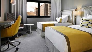 A hotel room with two beds, a desk, a chair, and a window with a city view.