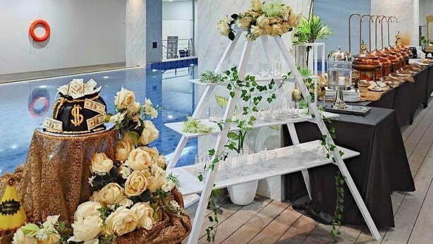 Une salle de réception avec un présentoir blanc à plusieurs niveaux, un gâteau décoré de billets d’un dollar et une rangée de tables ornées de verres.