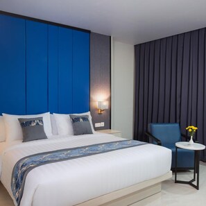 A hotel room with a large bed, a blue accent wall, a wall-mounted light, a blue armchair, and a small table with a vase of flowers. - Horison Ultima Entrop Papua (Jayapura)