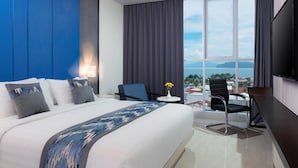 Une chambre d’hôtel avec un grand lit, un bureau, une chaise, une petite table avec un vase et une grande fenêtre donnant sur un beau paysage.