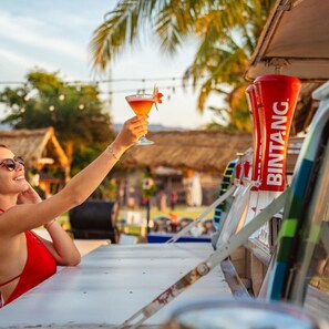 Una persona sosteniendo un vaso de cóctel con una bebida tropical, una licuadora marca Bintang y un entorno rústico al aire libre con palmeras y una estructura de techo de paja.