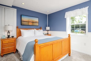 Un dormitorio con cama de madera, ropa de cama blanca, mesita de noche con lámpara, velador con reloj y ventana con vistas a la vegetación.