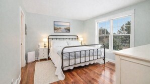 Quarto com estrutura de cama em metal, roupa de cama branca, mesa de cabeceira com abajur, quadro na parede e uma grande janela com vista para árvores.