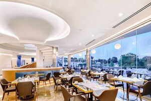 Un restaurante moderno con una barra central, varias mesas y grandes ventanales que ofrecen vistas al exterior.