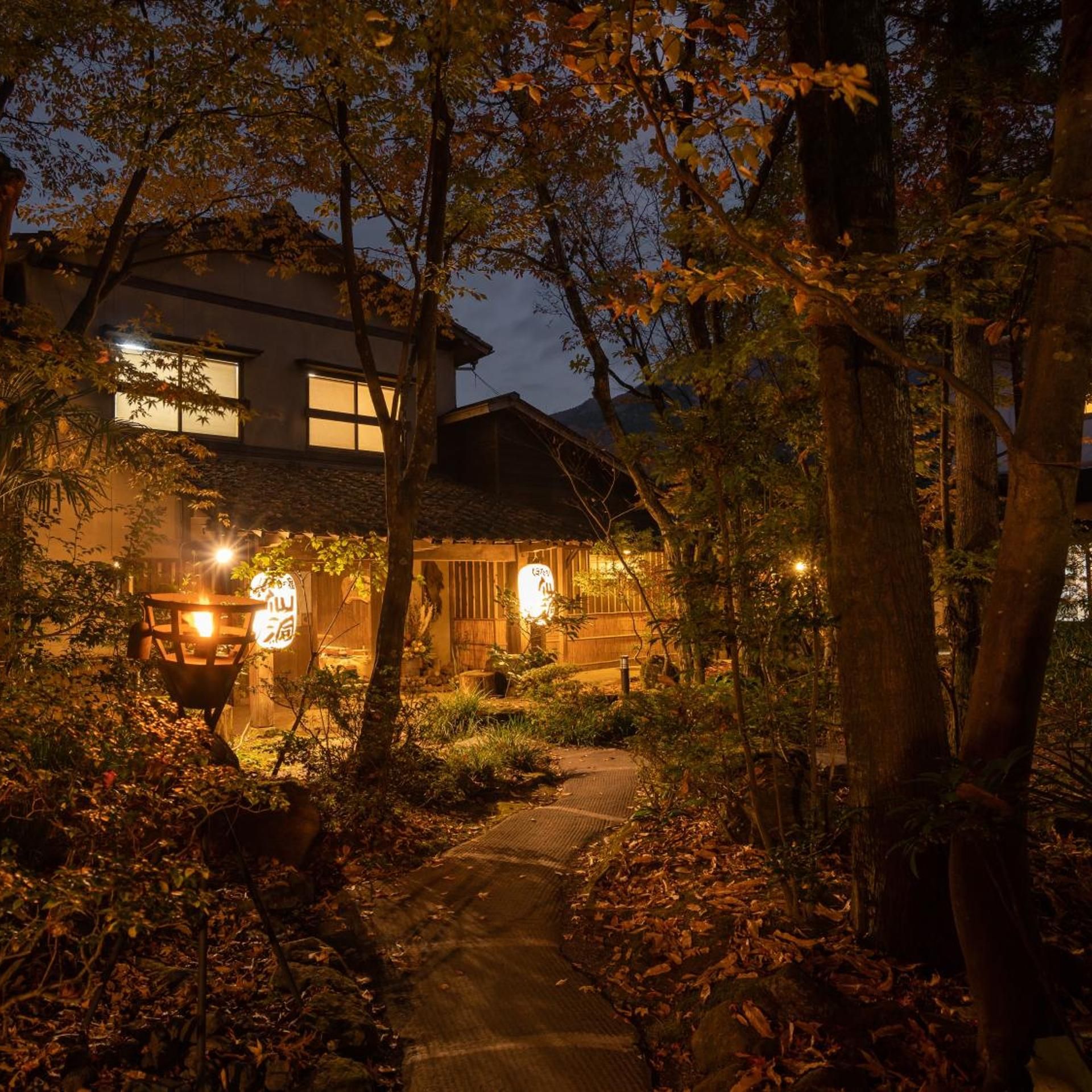 A traditional Japanese house at dusk with a pathway leading to the entrance, surrounded by trees and illuminated by warm lighting.