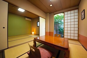 Ein Raum mit Tatami-Matten, einem Holztisch und -stühlen, einer verschiebbaren Shoji-Schirmwand und Blick ins Grüne.