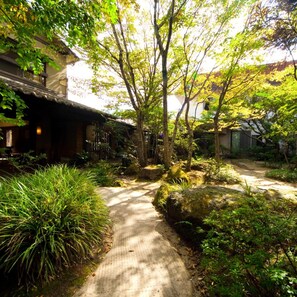 A pathway leading through a garden with trees and shrubs, a building with a traditional design, and a clear sky. - Hotarunoyado Sendou (Yufu)