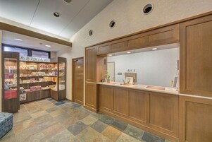 Un área de recepción con mostrador de madera, zona de estar con piso estampado, y una exhibición de libros y revistas.