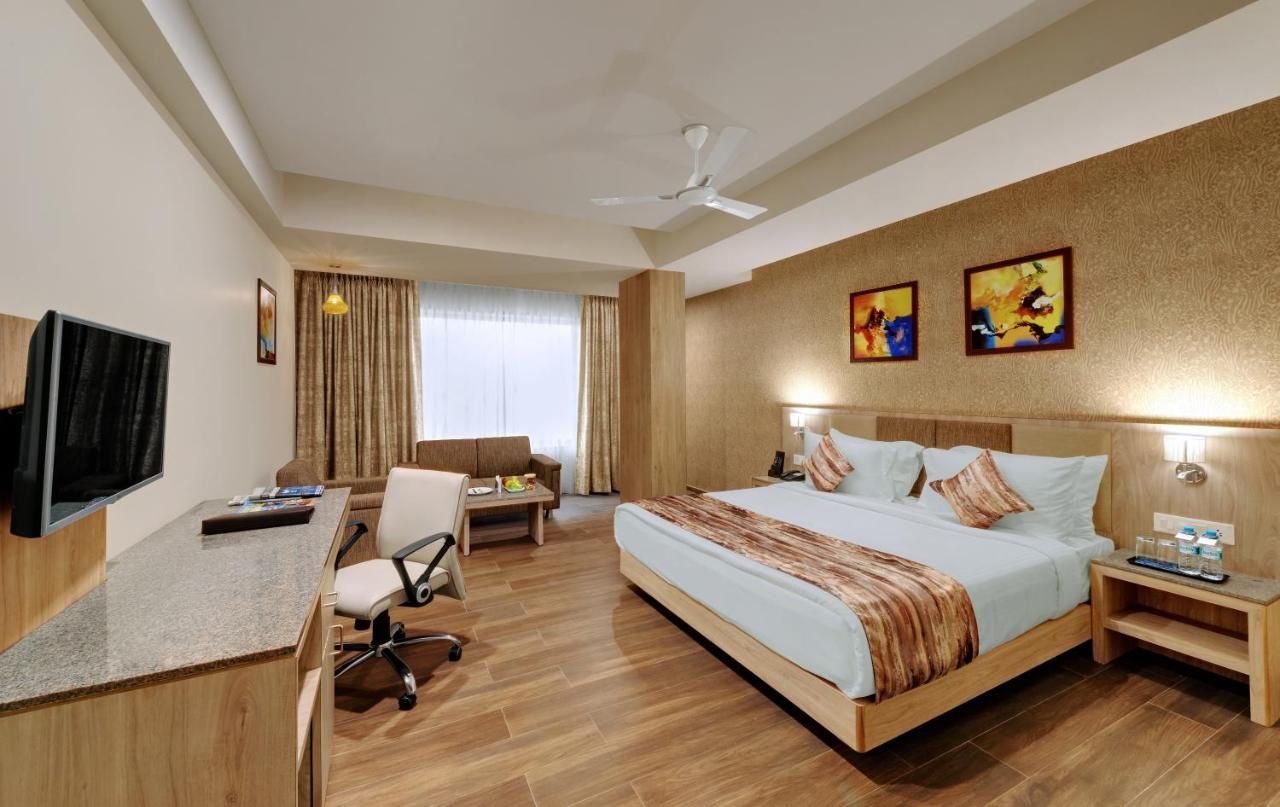 Una habitación de hotel con cama, escritorio, silla, televisor, obras de arte y ventilador de techo.