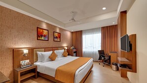 A hotel room with a bed, bedside table, lamp, artwork, curtains, chair, TV, and a ceiling fan. - Anaya Beacon Hotel (Jamnagar)