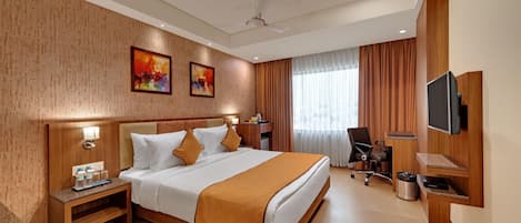 A hotel room with a bed, bedside table, lamp, artwork, curtains, chair, TV, and a ceiling fan.