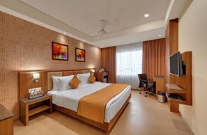 A hotel room with a bed, bedside table, lamp, artwork, curtains, chair, TV, and a ceiling fan. - Anaya Beacon Hotel (Jamnagar)