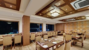 A hotel dining room with multiple tables and chairs, large windows, and a patterned wall. - Anaya Beacon Hotel (Jamnagar)