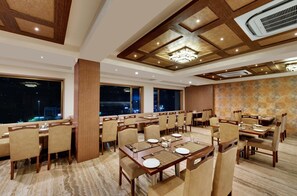 A hotel dining room with multiple tables and chairs, large windows, and a patterned wall. - Anaya Beacon Hotel (Jamnagar)