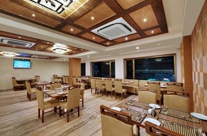 A hotel dining area with multiple tables and chairs, a large screen, and a view of the city at night. - Anaya Beacon Hotel (Jamnagar)