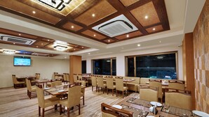 A hotel dining area with multiple tables and chairs, a large screen, and a view of the city at night. - Anaya Beacon Hotel (Jamnagar)
