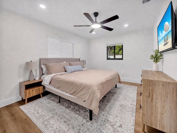 A bedroom with a bed, nightstands, a ceiling fan, a window, a small plant, and a TV.