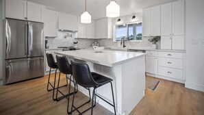 Une cuisine moderne avec des meubles blancs, un réfrigérateur en acier inoxydable, un îlot central doté de tabourets, des luminaires suspendus et une fenêtre donnant sur un paysage urbain.