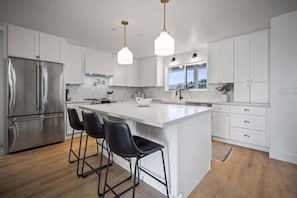 A modern kitchen with white cabinetry, a stainless steel refrigerator, a central island with bar stools, pendant lighting, and a window overlooking a cityscape. - Aether - Sparkling Pool & Games - Mins to Old Town (Scottsdale)