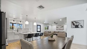 Une cuisine moderne avec des meubles blancs, un coin repas doté d’une grande table et de chaises, et un salon avec un canapé et un tableau au mur.
