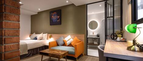 A modern hotel room with a bed, a sofa, a desk, and a bathroom visible through a glass door.