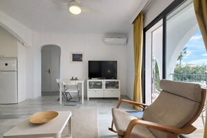 Ein modernes Wohnzimmer mit einem Flachbildfernseher, einem Essbereich mit weißem Tisch und Stühlen sowie ein Balkon mit gelben Vorhängen.