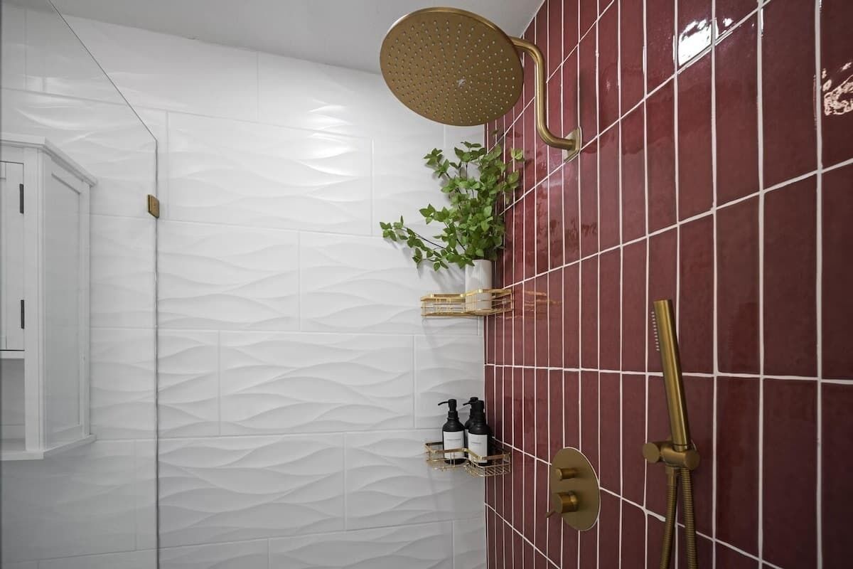 A bathroom with a white textured wall, a red tiled wall, a gold showerhead, a gold handheld showerhead, and a plant in a gold planter.