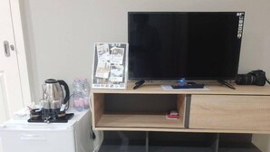 A small kitchenette with a coffee maker, a kettle, and a refrigerator. There is a flat-screen TV on a wooden stand with a camera and a magazine on it. - Iresidence Hotel Pathumthani (Khlong Luang)