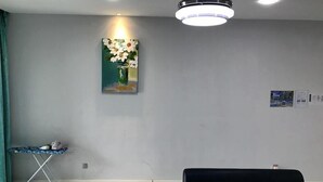 A living room with a black sofa, a small table with an ironing board, a painting of flowers, and a ceiling light fixture. - Sunset & Seaview Vacation Condo City Centre @ Imago Shopping Mall (Kota Kinabalu)