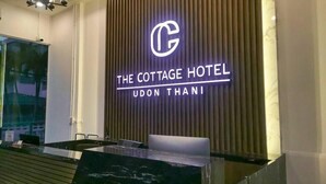 Un comptoir d’accueil au design moderne, un logo placé au-dessus du nom « The Cottage Hotel Udon Thani », et un panneau vertical avec des lattes verticales en arrière-plan.