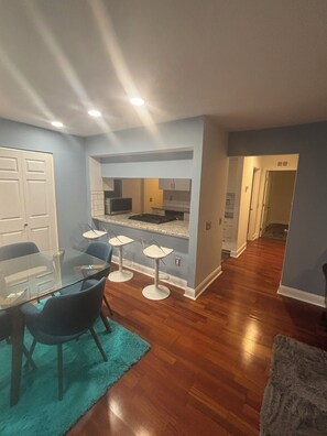 A modern kitchen with a central island, a dining area with a glass table and chairs, and a hallway leading to another room.
