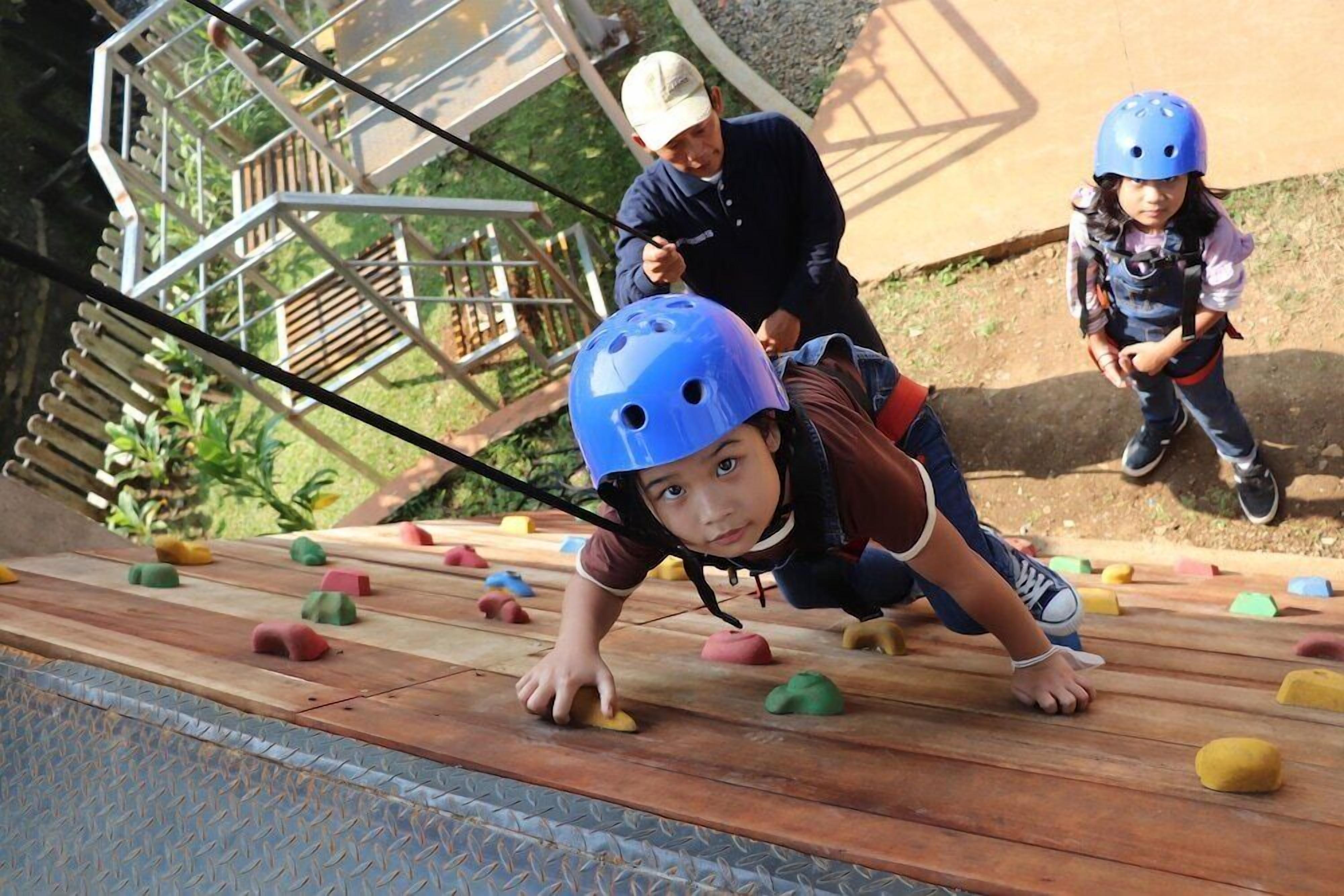 Un enfant escalade un parcours d’obstacles en bois, composé de blocs de mousse colorés. Il porte un casque bleu et un harnais de sécurité, et est surveillé par un adulte.