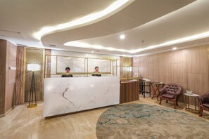 A reception desk with a marble top, a large mirror, and a modern ceiling design. There are leather chairs and a small table with a lamp. - Bigland Hotel Bogor (Bogor)