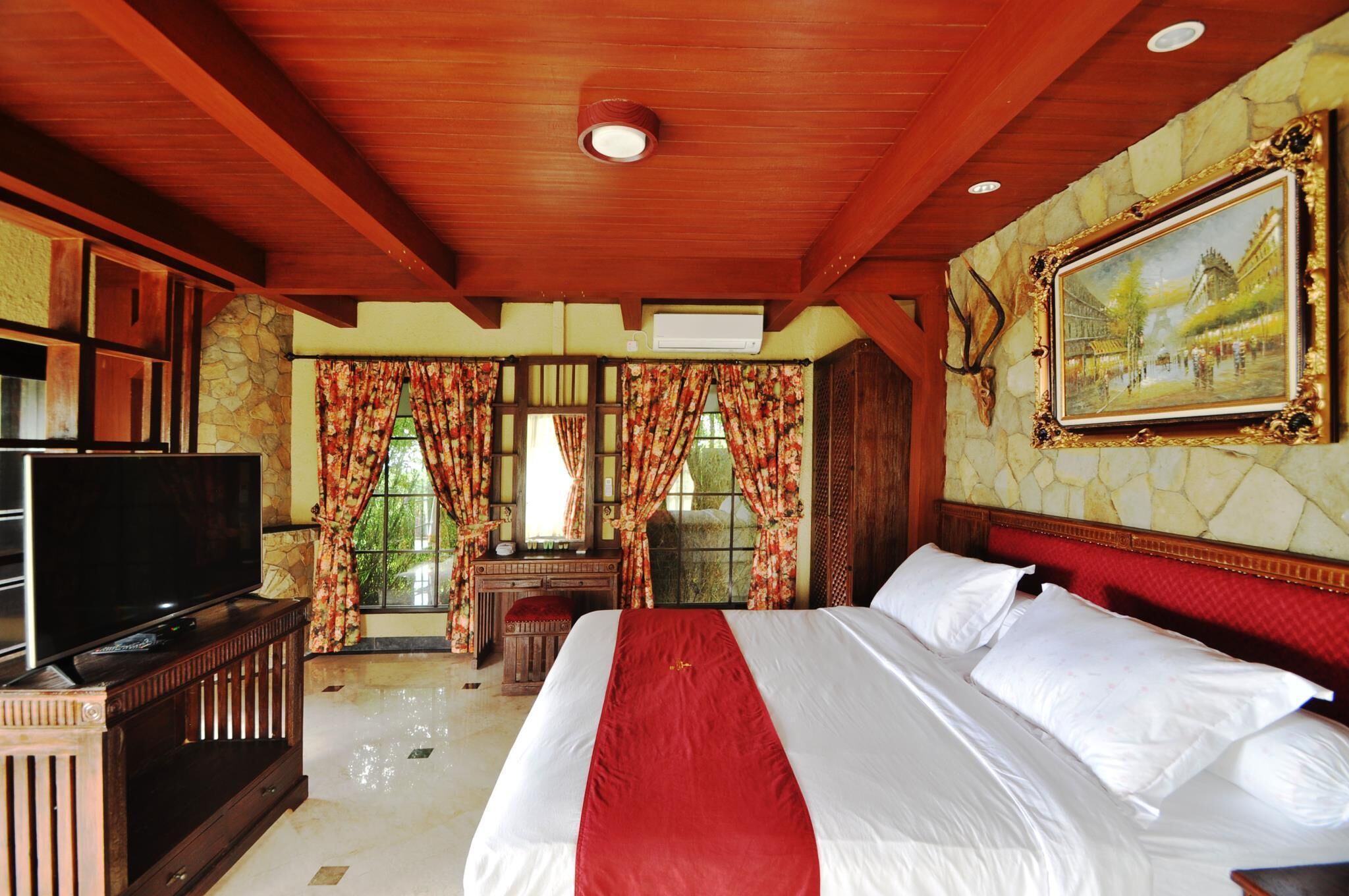 A spacious bedroom with a large bed, a television on a wooden stand, and a stone wall with a framed painting.