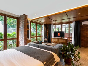 Une chambre d’hôtel moderne dotée d’un grand lit, d’un coin salon, d’une télévision et offrant une vue sur la verdure grâce à de grandes fenêtres.