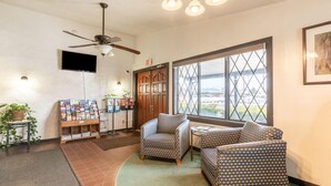A lobby with a ceiling fan, a TV mounted on the wall, a magazine rack, a wooden door, and a large window with a view of the outside.