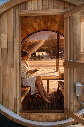 Una sauna de madera con un banco y un asiento, una persona leyendo un libro, y vista a un paisaje nevado.