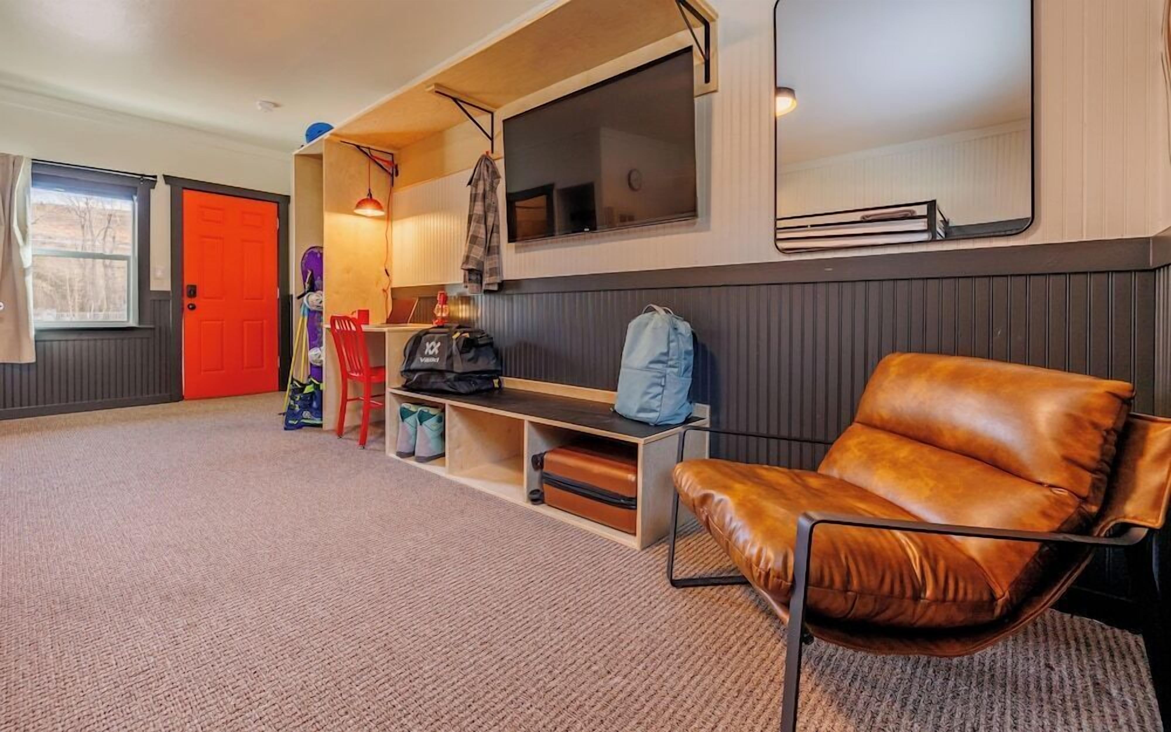 A room with a brown leather chair, a red door, a desk with a lamp, and a large window.