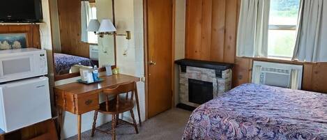 A room with a bed, a desk with a chair, a television, a microwave, a fireplace, and a window with curtains.