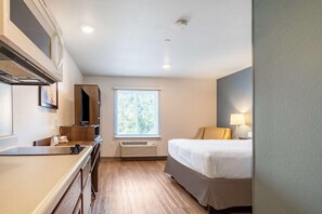 A hotel room with a kitchenette, a bed, a window with blinds, and a lamp. - Woodspring Suites Portland North Gresham (Portland)