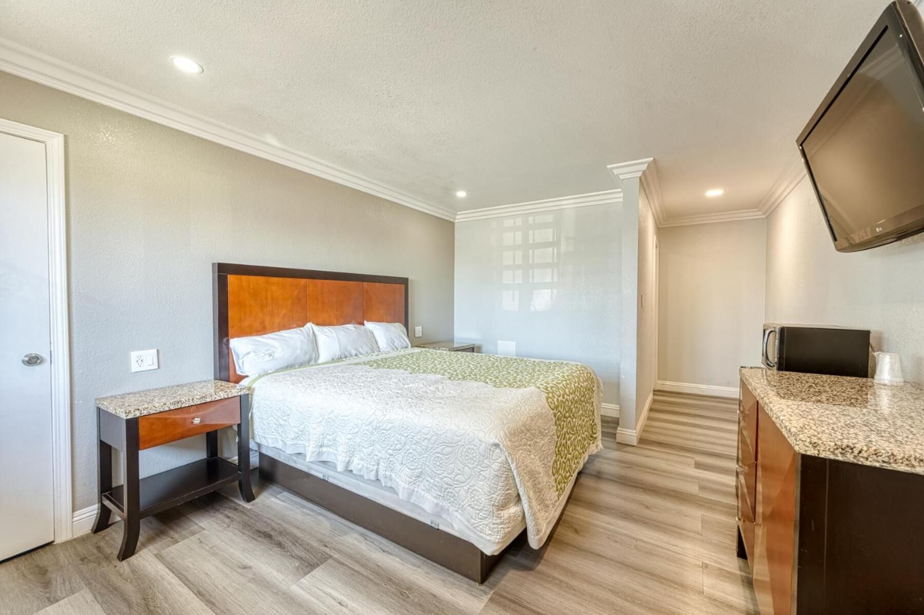 A hotel room with a bed, bedside table, TV, and a kitchenette area.