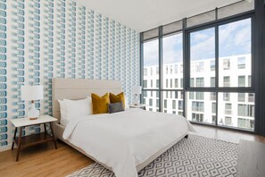Una habitación de hotel moderna con una cama grande, una mesita de noche, un tapete estampado y vistas a la ciudad a través de amplias ventanas.