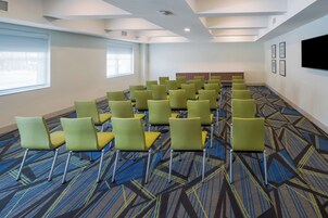 Un salón de conferencias con filas de sillas verdes, una alfombra estampada y un televisor de pantalla plana en la pared.