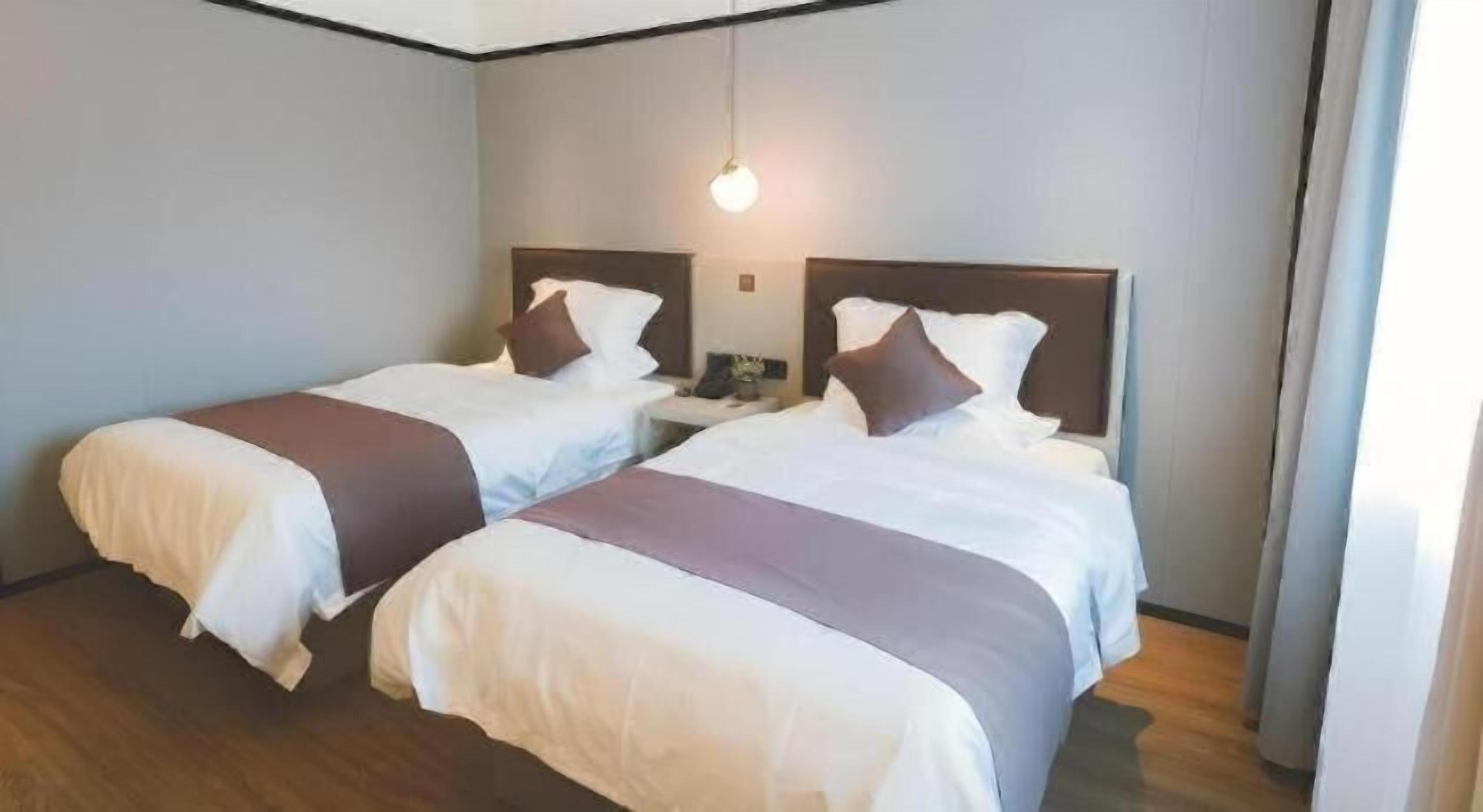 A hotel room with two beds, a wooden floor, a headboard with a light fixture, and a nightstand with a lamp.